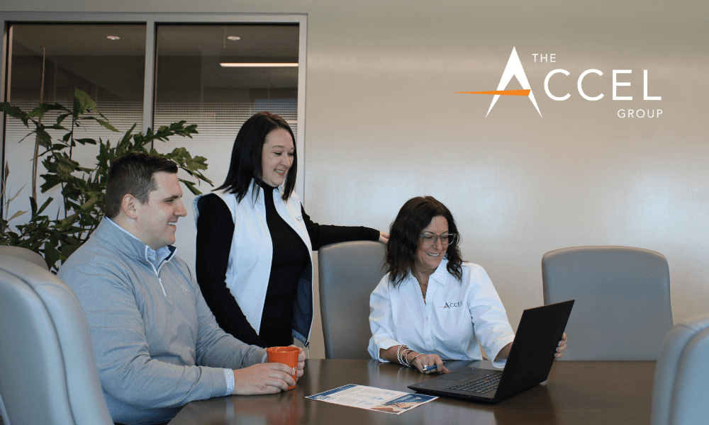 Photo of two women and a man in a boardroom smiling at a laptop with The Accel Group logo
